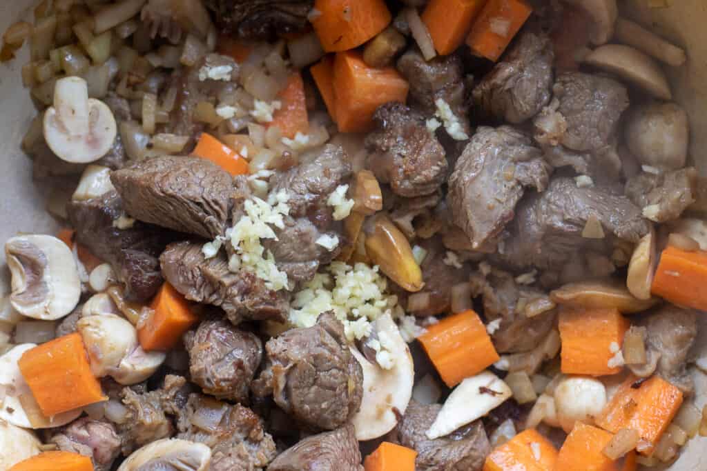 Browned beef is cooking with carrots, garlic and mushrooms.