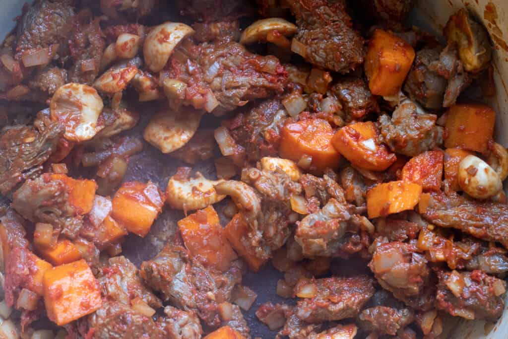 Browned beef cooking with onions, carrots, mushrooms, garlic, tomato paste, and flour.