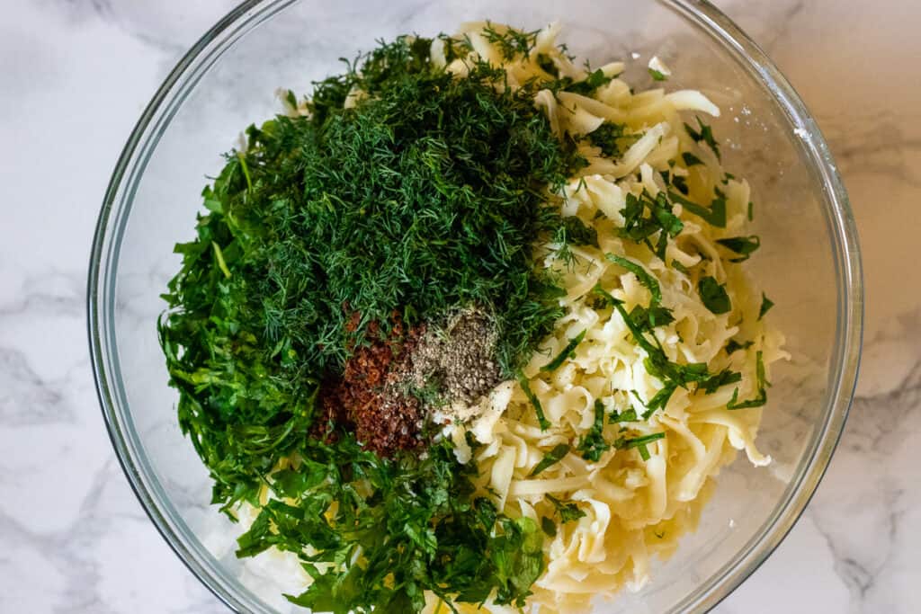 Grated cheese, chopped herbs, chili flakes, and black pepper in a glass bowl before mixing.