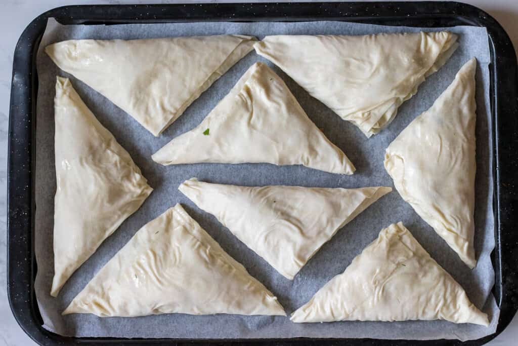 Unbaked cheese borek triangles arranged on a parchment-lined baking tray.