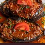 Two stuffed eggplants in tomato sauce on a white plate, topped with tomato slices and parsley.