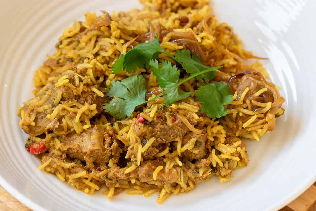 A plate of biryani loaded with aromatic yellow rice, tender pieces of marinated meat, caramelized onions, and garnished with fresh cilantro leaves. The dish is rich in spices and appears flavorful, served on a white plate.