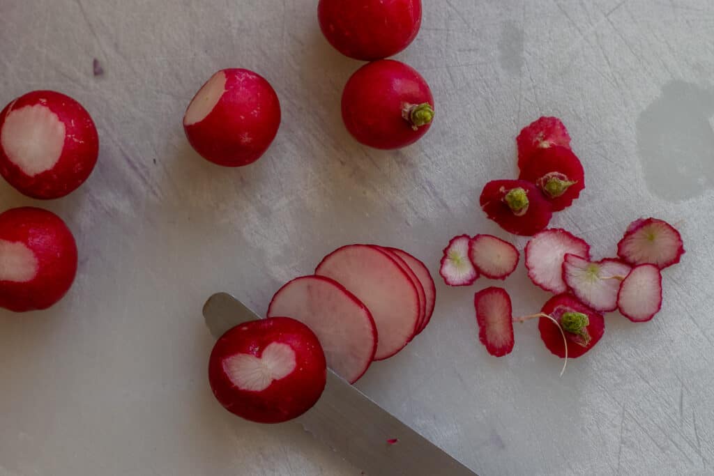 Radishes being thinly sliced on a cutting board.