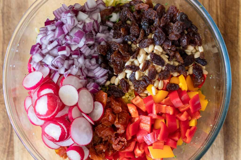 Orzo pasta salad in a glass bowl topped with red onion, radishes, sun-dried tomatoes, peppers, pine nuts, and sultanas before mixing.