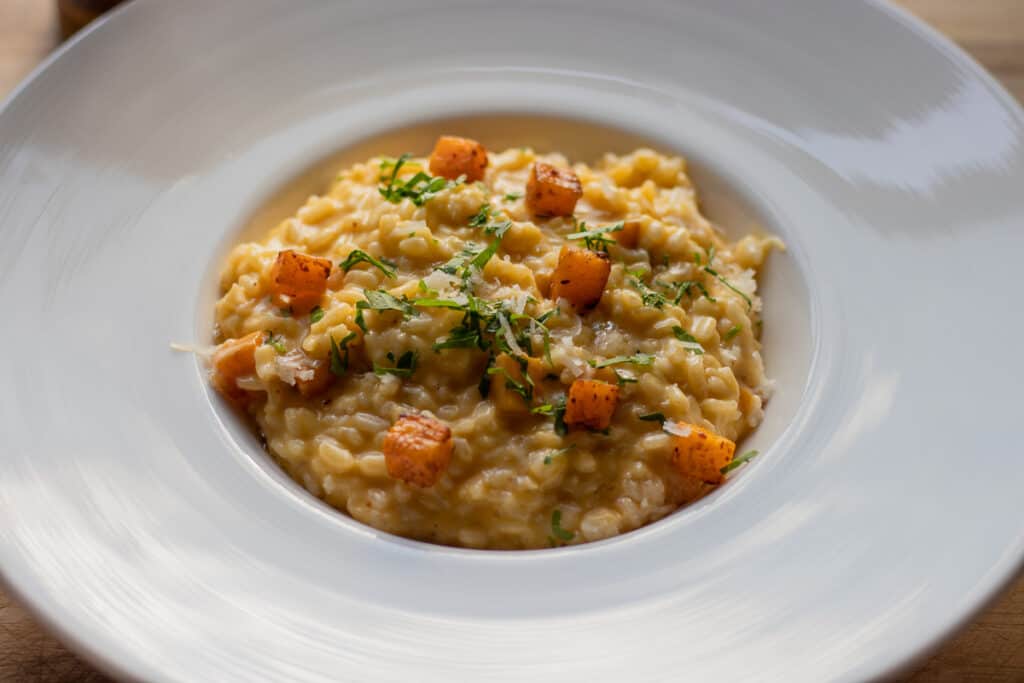 A white plate of creamy risotto garnished with small cubes of butternut squash and sprinkled with finely chopped green herbs. The dish has a rich, golden-yellow color and appears smooth and hearty.
