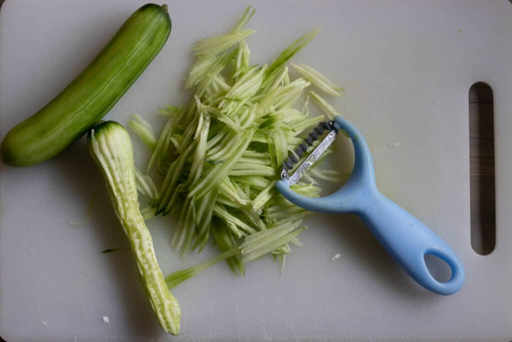 Mini cucumber being cut into thin matchsticks with a julienne peeler.