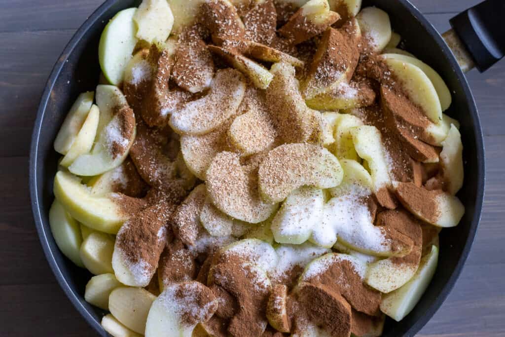 Sliced apples are placed in a pan with cinnamon, nutmeg, and sugar.