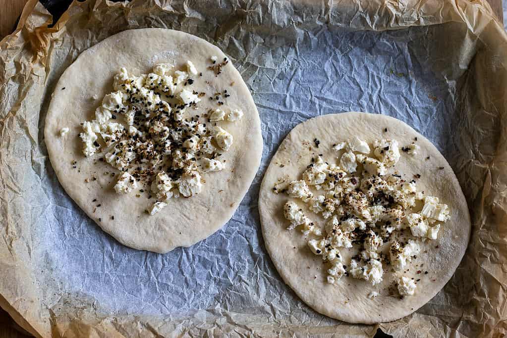 Crumbled feta and nigella seeds on two dough circles.