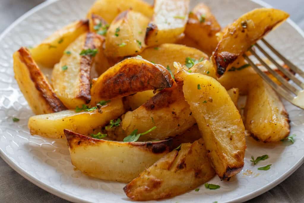 A close-up of a plate of golden, roasted potato wedges garnished with chopped parsley. The potatoes have a crispy exterior and are arranged on a white plate with a fork visible on the right side.