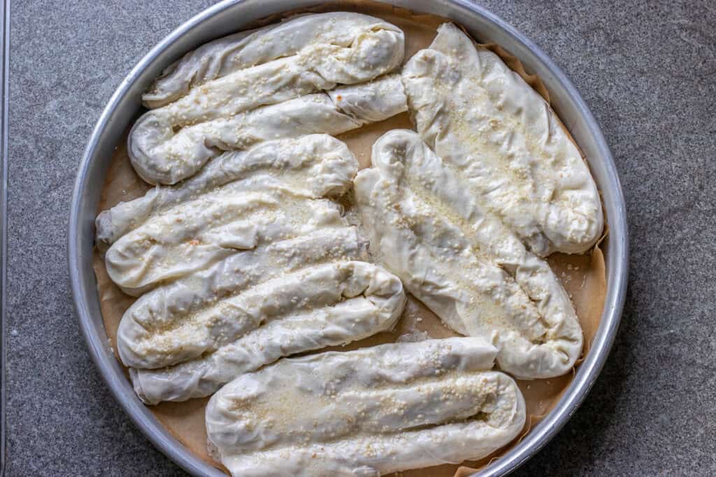 Six shaped kol boregi arranged in a round baking pan.