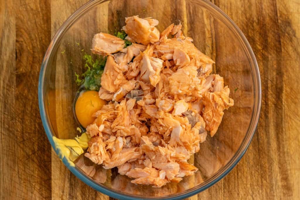 Flaked cooked salmon added to the rest of the patty ingredients in a glass bowl.