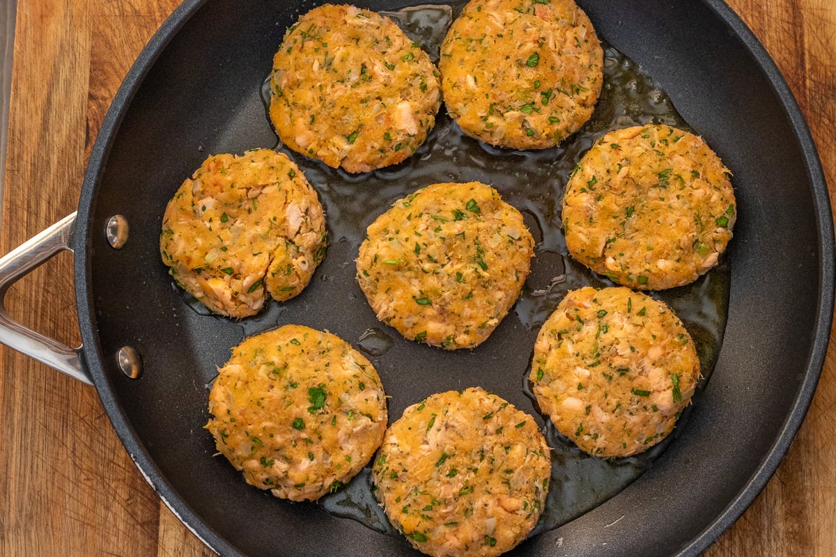 Salmon patties frying in oil in a black nonstick pan.