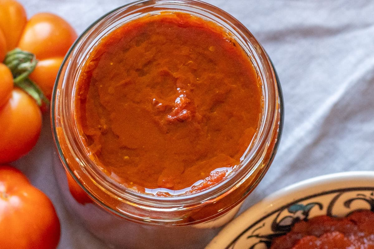 An open jar of homemade tomato paste with fresh tomatoes beside it.